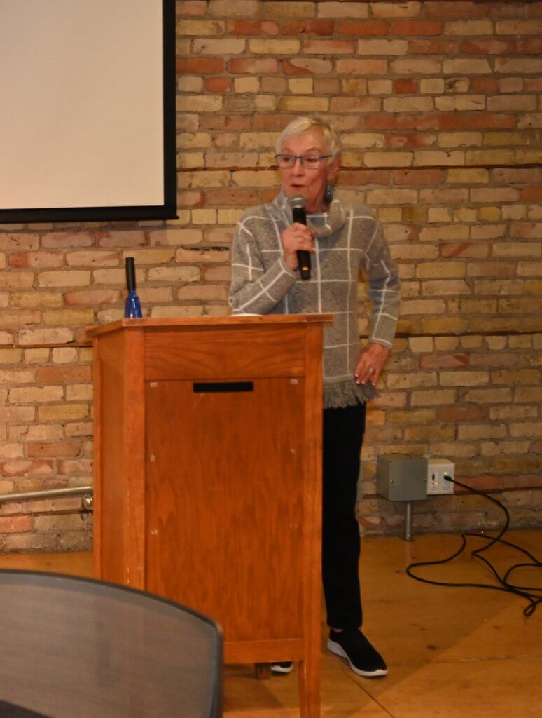 Bridges to Learning Founder holds the mic by the podium and thanks the guests. 2022 Wine Tasting to raise funds for poor children in Vietnam. Nonprofit.