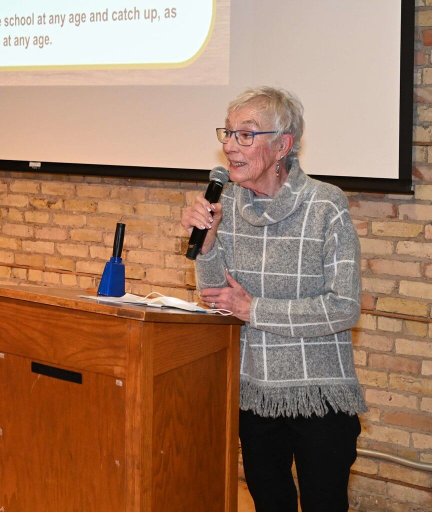 Bridges to Learning Founder holds the mic by the podium and thanks the guests. 2022 Wine Tasting to raise funds for poor children in Vietnam. Nonprofit.
